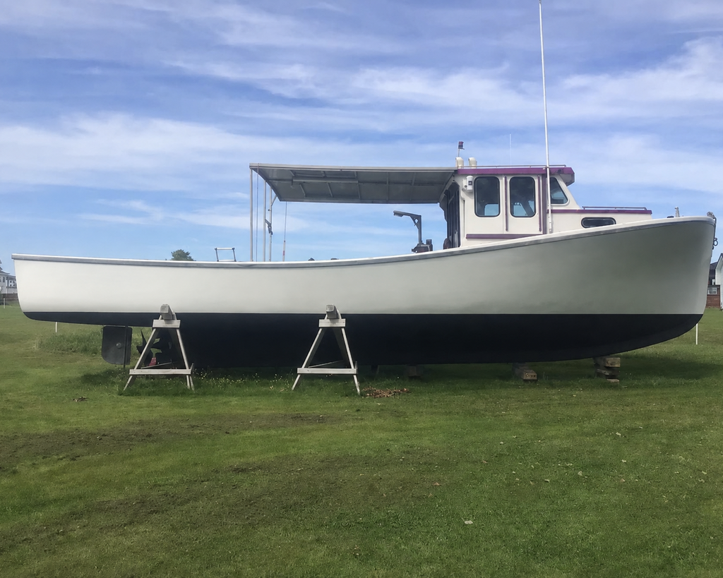 A white and black boat with a purple cabin trim sits on metal stands on a grassy lawn under a blue sky with clouds.