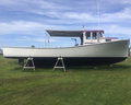 A white and black boat with a purple cabin trim sits on metal stands on a grassy lawn under a blue sky with clouds.