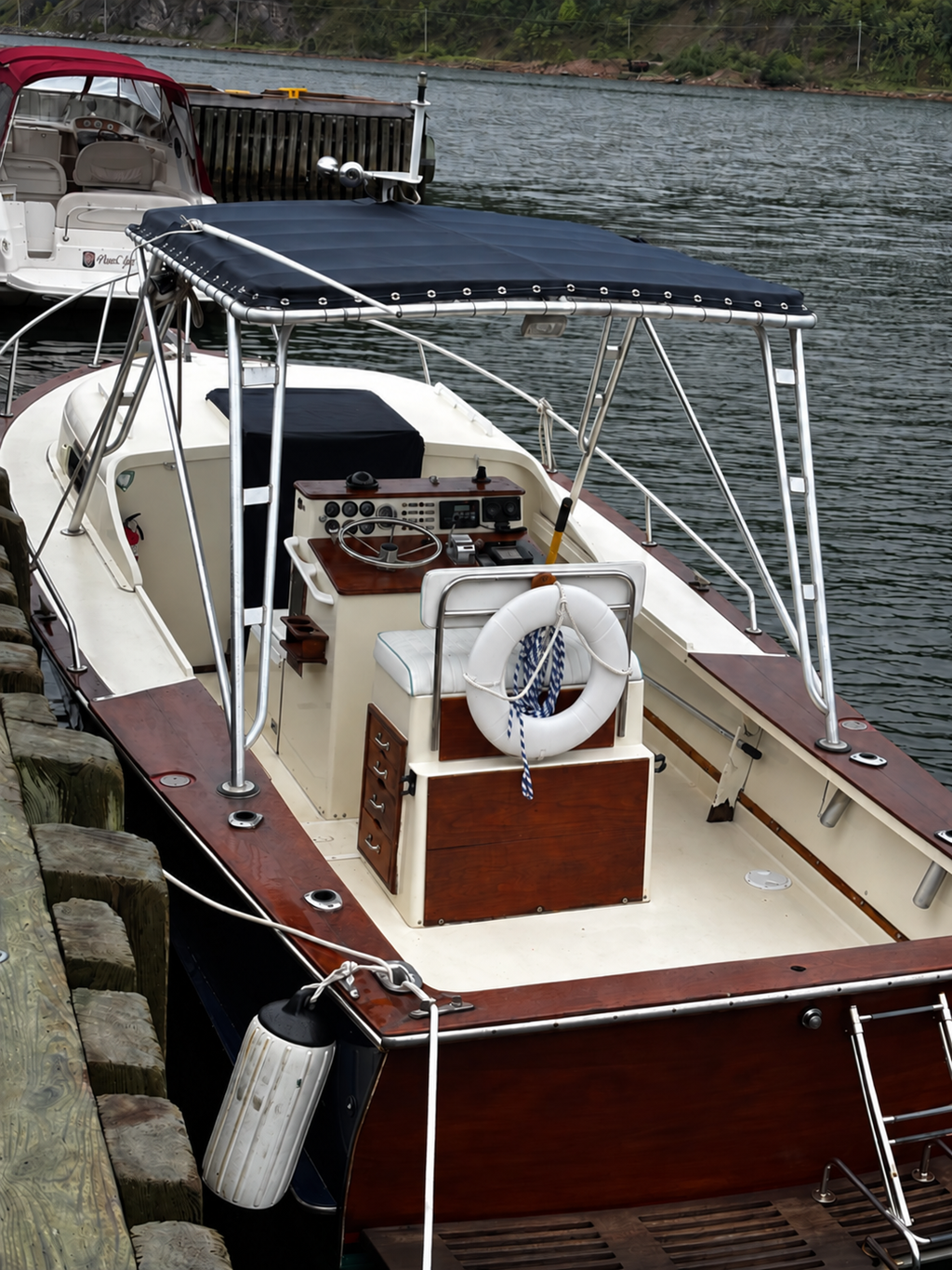 A classic wooden motorboat docked at a pier with water and trees in the background. The boat features a cream-colored interior, dark reddish-brown wood trim, a dark blue canvas bimini top, a white life preserver, and a wooden helm console.