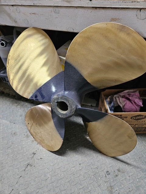 A four-blade boat propeller with brass-colored blades and a dark gray metal hub, photographed on a concrete floor in a workshop setting.