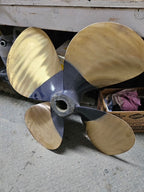 A four-blade boat propeller with brass-colored blades and a dark gray metal hub, photographed on a concrete floor in a workshop setting.