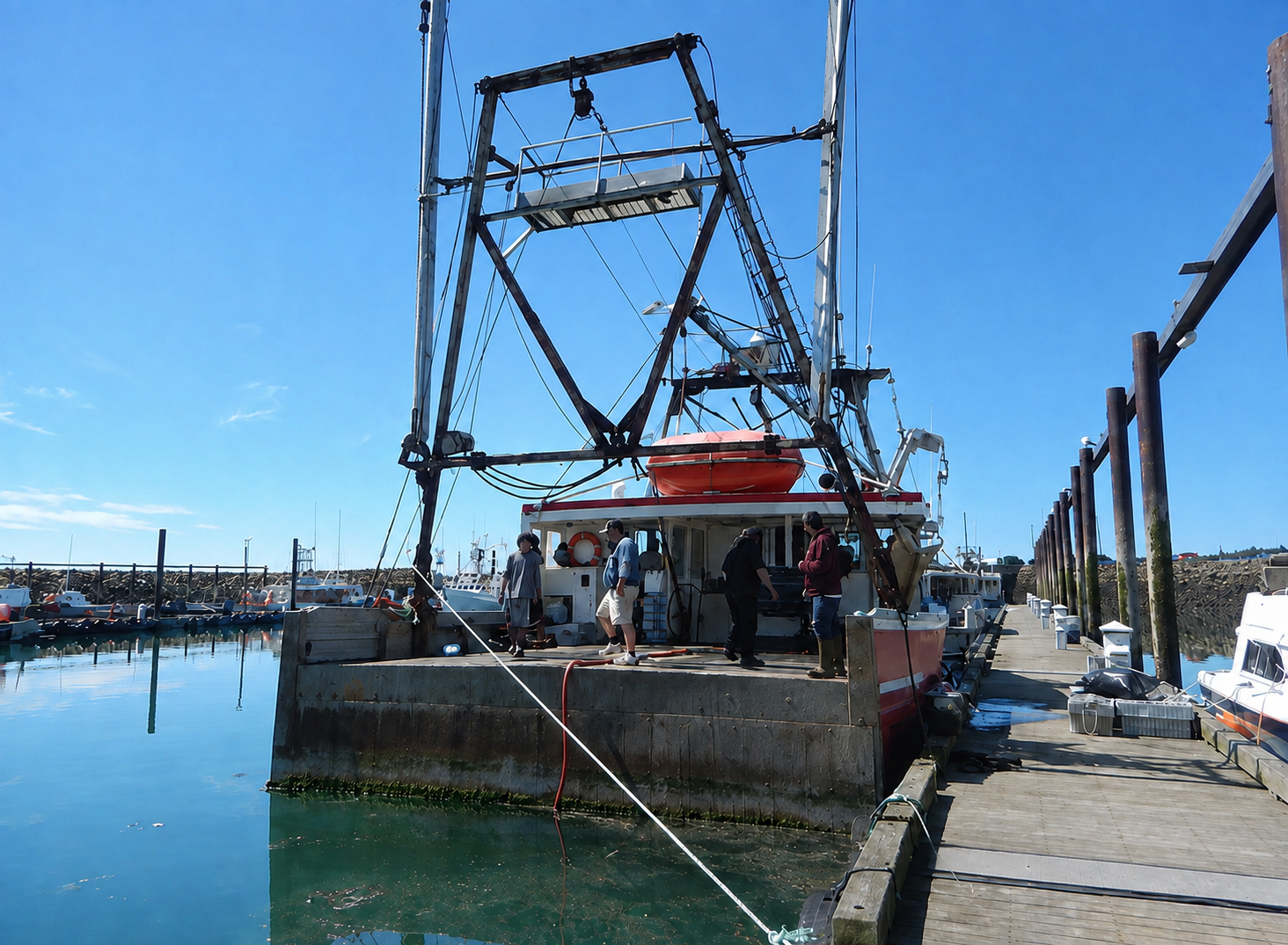 A large fishing vessel docked at a harbor pier under a clear blue sky, with several people standing on the deck and calm water reflecting around the boat.