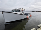 A white fishing boat with a black hull stripe and cabin, floating on calm water under an overcast sky. Orange buoys are attached to the side of the boat, and a dock is visible in the bottom right corner.