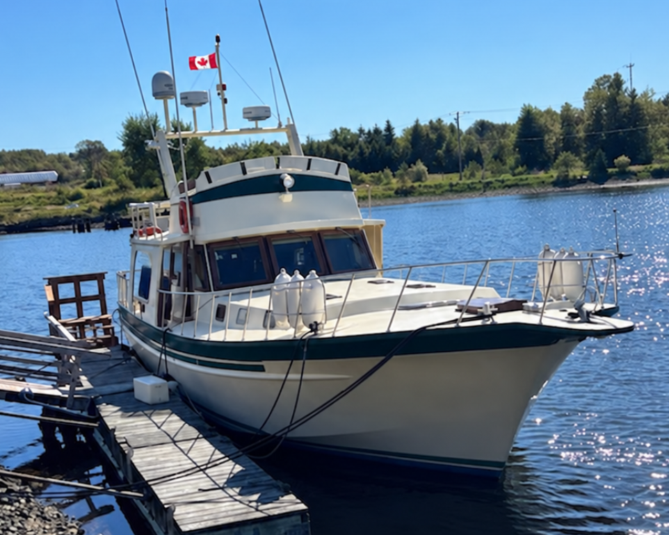 A white and dark green fishing boat with a Canadian flag on the mast, docked at a wooden pier on a sunny day with blue water and trees in the background.