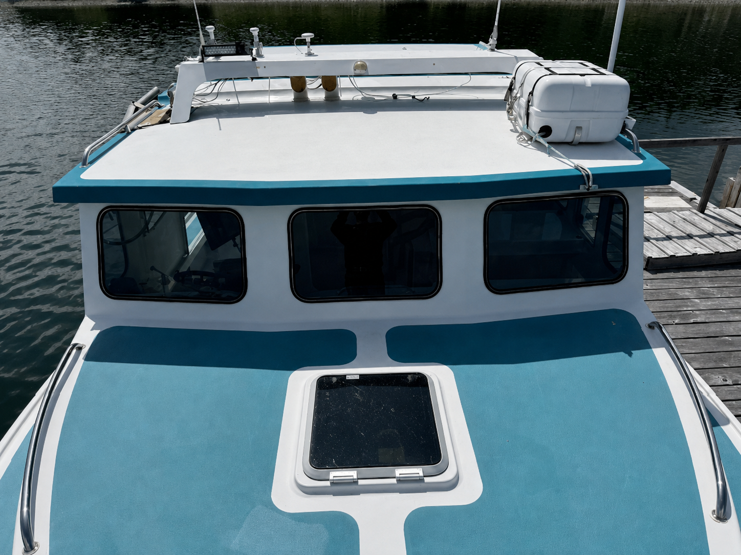 Close-up of the front cabin and deck of a blue and white boat docked at a wooden pier with water visible in the background.