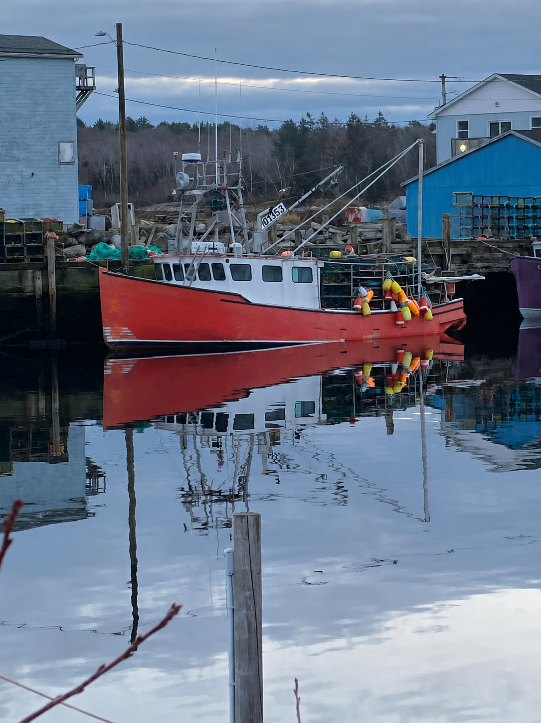 A red and white fishing boat docked in calm water, with its reflection clearly visible on the surface. Small buildings, utility poles, and trees line the background under an overcast sky.