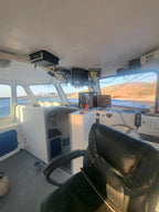 The helm station of a boat cabin with a black leather captain's chair in the foreground, white control consoles, and multiple monitors mounted on the ceiling and dashboard, with water and a shoreline visible through the windows.