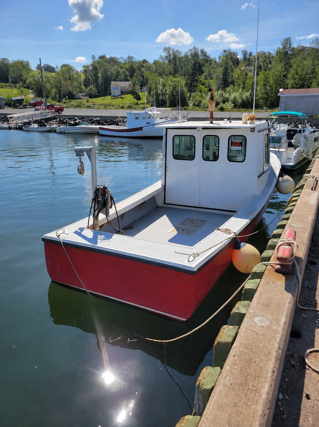 A small red and white fishing boat docked at a harbor under a blue sky with clouds, surrounded by other boats and green trees in the background.