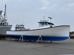 A white and blue fishing boat on wooden supports in an outdoor boatyard, with other boats and a cloudy sky in the background.