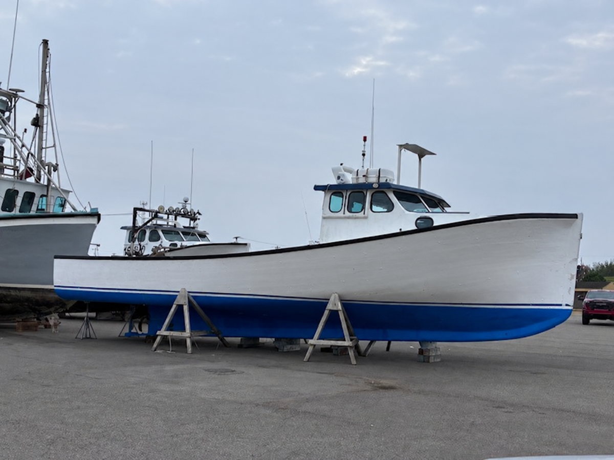 A white and blue fishing boat on wooden supports in an outdoor boatyard, with other boats and a cloudy sky in the background.
