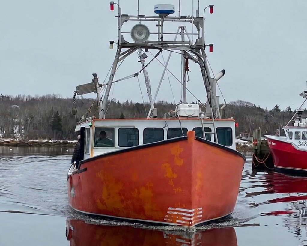 A red and white fishing boat with a tall metal rigging mast floats in calm water, showing its reflection on the surface. Another boat is partially visible on the right, and a tree-lined shore sits in the background under an overcast sky.