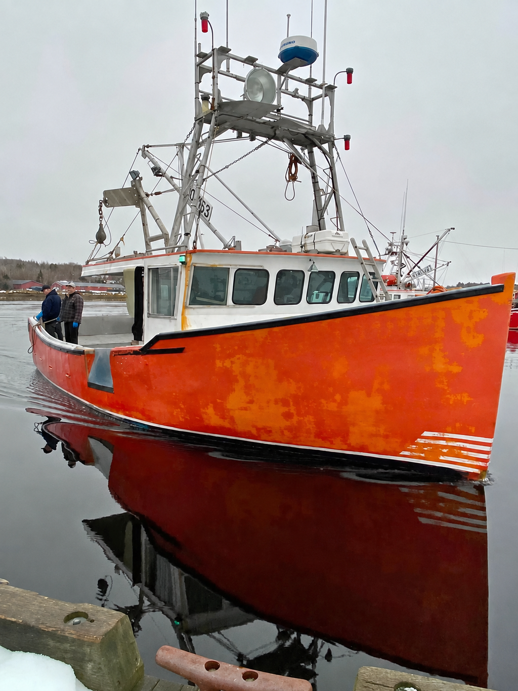 A red and white fishing boat docked in calm water under an overcast sky, with its reflection visible on the surface. A person is standing on the deck near the bow.