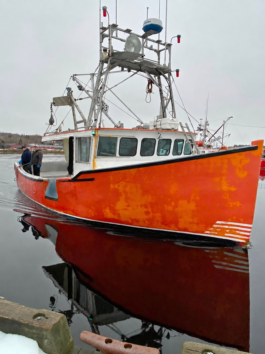 A red and white fishing boat docked in calm water under an overcast sky, with its reflection visible on the surface. A person is standing on the deck near the bow.