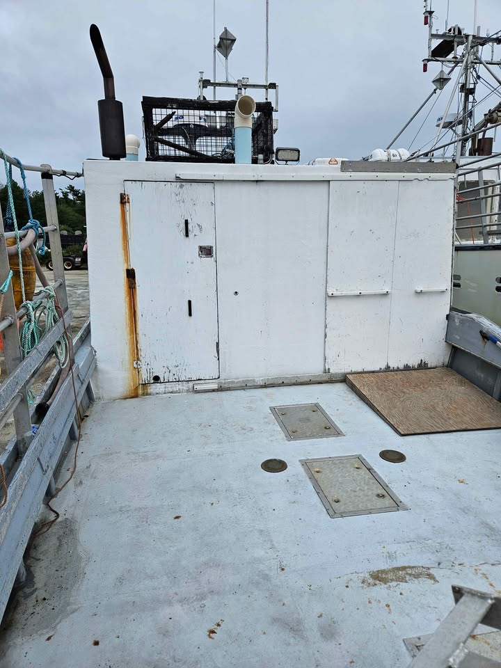 A white metal utility cabin on the deck of a fishing boat under an overcast sky, with metal railings, rigging, and deck hatch covers visible.