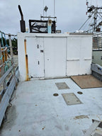 A white metal utility cabin on the deck of a fishing boat under an overcast sky, with metal railings, rigging, and deck hatch covers visible.