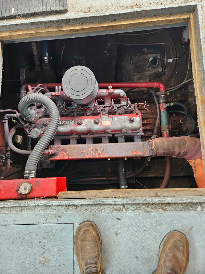 A close-up view of a red and gray industrial engine or generator unit inside a metal compartment, with a person's brown shoes visible at the bottom edge.