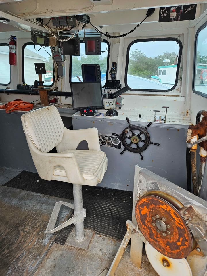 The interior of a boat's navigation deck featuring a white plastic captain's chair, a wooden ship's wheel, various electronic screens, and a weathered metal propeller in the foreground.