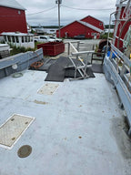The back deck of a boat with a light gray fiberglass floor, metal railings, black mats, and a wooden chair, with red barns and a dock in the background.