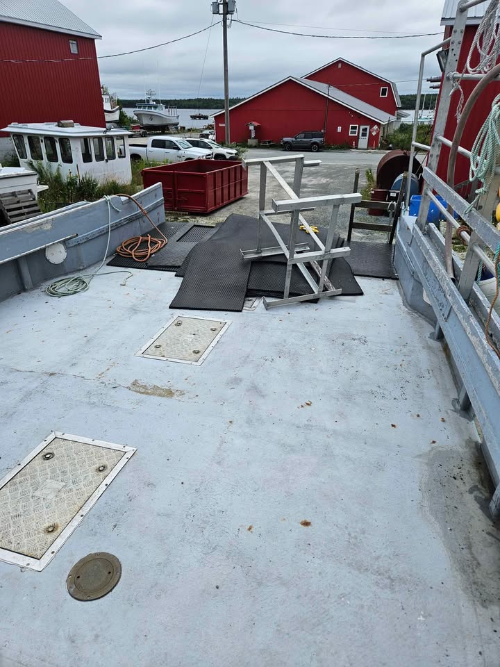 The back deck of a boat with a light gray fiberglass floor, metal railings, black mats, and a wooden chair, with red barns and a dock in the background.