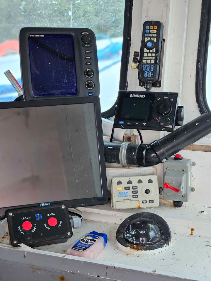 A collection of marine electronic devices on a weathered white dashboard inside a boat cabin, including a Garmin fish finder, a Simrad radar detector, a Tiltech steering wheel controller, and other navigational equipment.