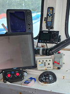 A collection of marine electronic devices on a weathered white dashboard inside a boat cabin, including a Garmin fish finder, a Simrad radar detector, a Tiltech steering wheel controller, and other navigational equipment.