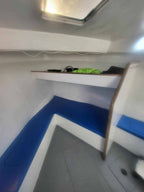 An interior corner of a boat cabin with white walls and floor, blue seating or cushioning along the walls, a shelf, and a small bright green item on the shelf.