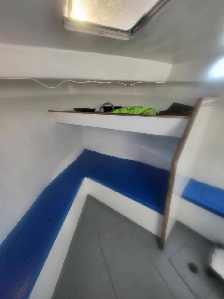 An interior corner of a boat cabin with white walls and floor, blue seating or cushioning along the walls, a shelf, and a small bright green item on the shelf.