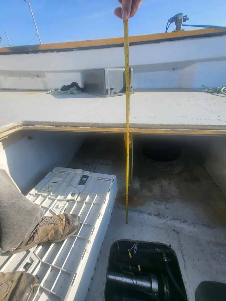 A hand holding a yellow tape measure inside the cockpit of a boat, showing the depth of the cockpit opening. The boat has a white hull with a wooden trim, and various boat fixtures and a cooler are visible.