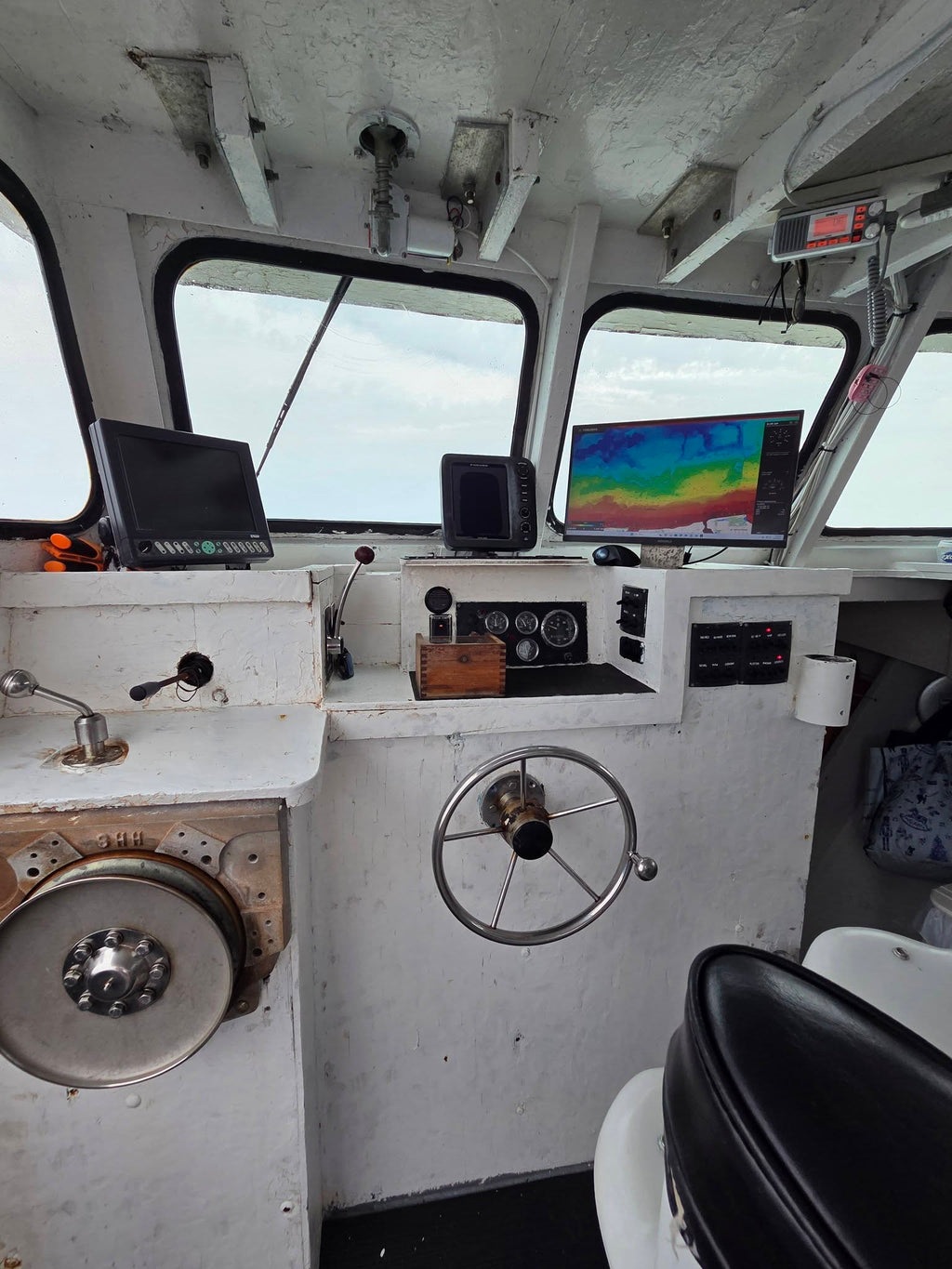 The interior of a boat's navigation deck featuring a white control console with a large metal steering wheel, various electronic screens, gauges, and a porthole window showing the open water outside.