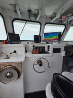 The interior of a boat's navigation deck featuring a white control console with a large metal steering wheel, various electronic screens, gauges, and a porthole window showing the open water outside.