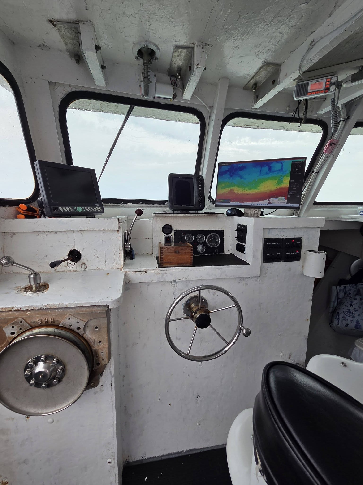 The interior of a boat's navigation deck featuring a white control console with a large metal steering wheel, various electronic screens, gauges, and a porthole window showing the open water outside.