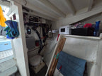 A cluttered interior space with white walls and ceiling beams, containing a microwave on a shelf, a blue quilted cushion, gloves hanging on the left, and various cables and equipment in the background.