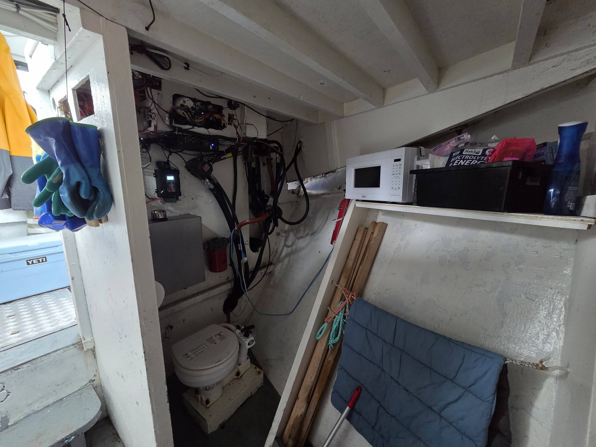 A cluttered interior space with white walls and ceiling beams, containing a microwave on a shelf, a blue quilted cushion, gloves hanging on the left, and various cables and equipment in the background.