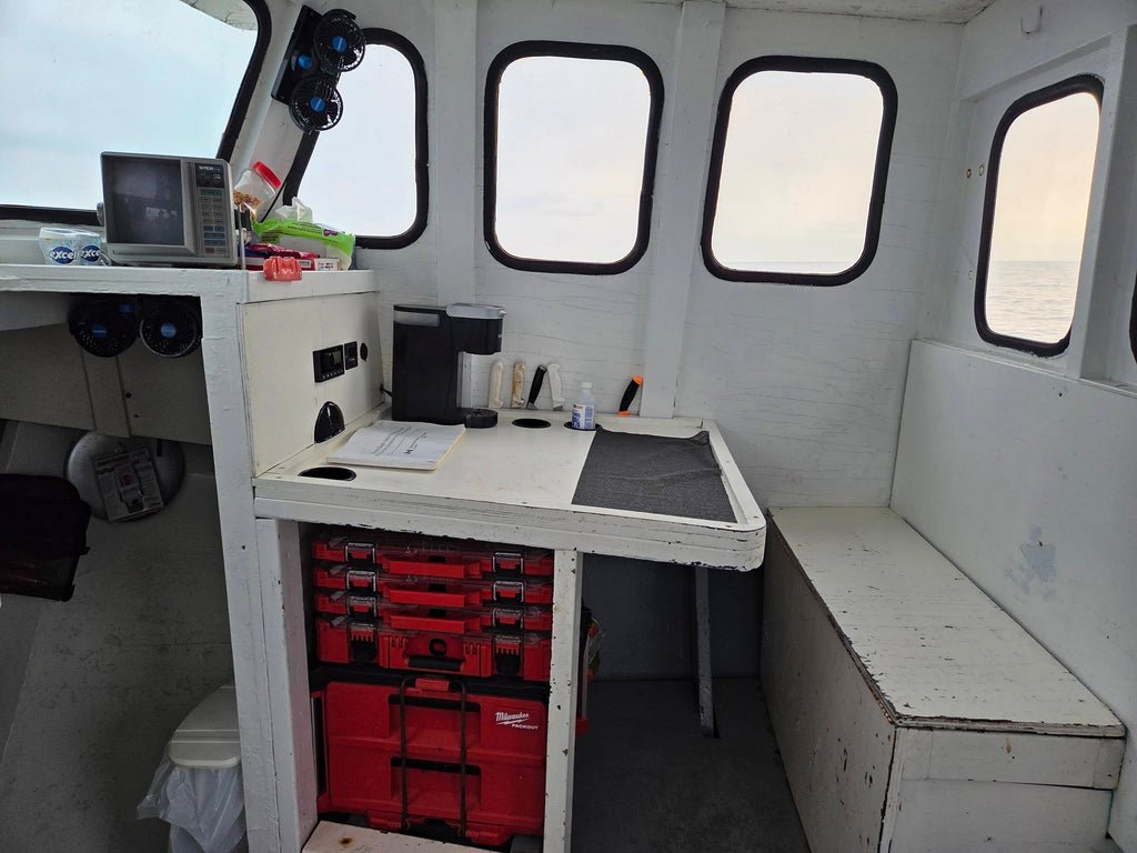 The interior of a boat cabin with a white wooden table, red storage toolboxes underneath, a microwave, and several windows looking out onto the water.