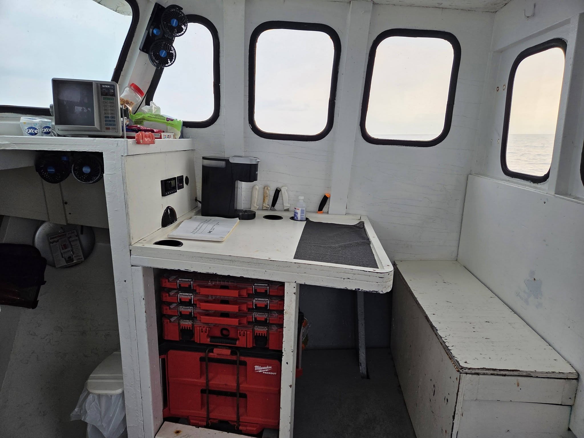 The interior of a boat cabin with a white wooden table, red storage toolboxes underneath, a microwave, and several windows looking out onto the water.