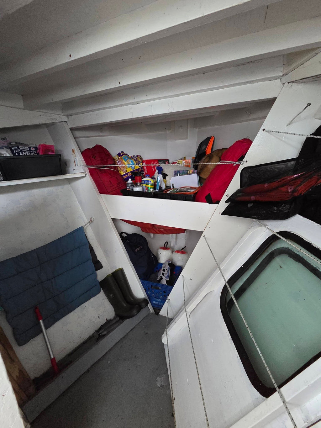A narrow, cluttered interior space with white walls and ceiling beams, containing shelves, a blue quilted mat, a broom, and various packed items including red and orange bags and boxes.
