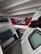 A narrow, cluttered interior space with white walls and ceiling beams, containing shelves, a blue quilted mat, a broom, and various packed items including red and orange bags and boxes.
