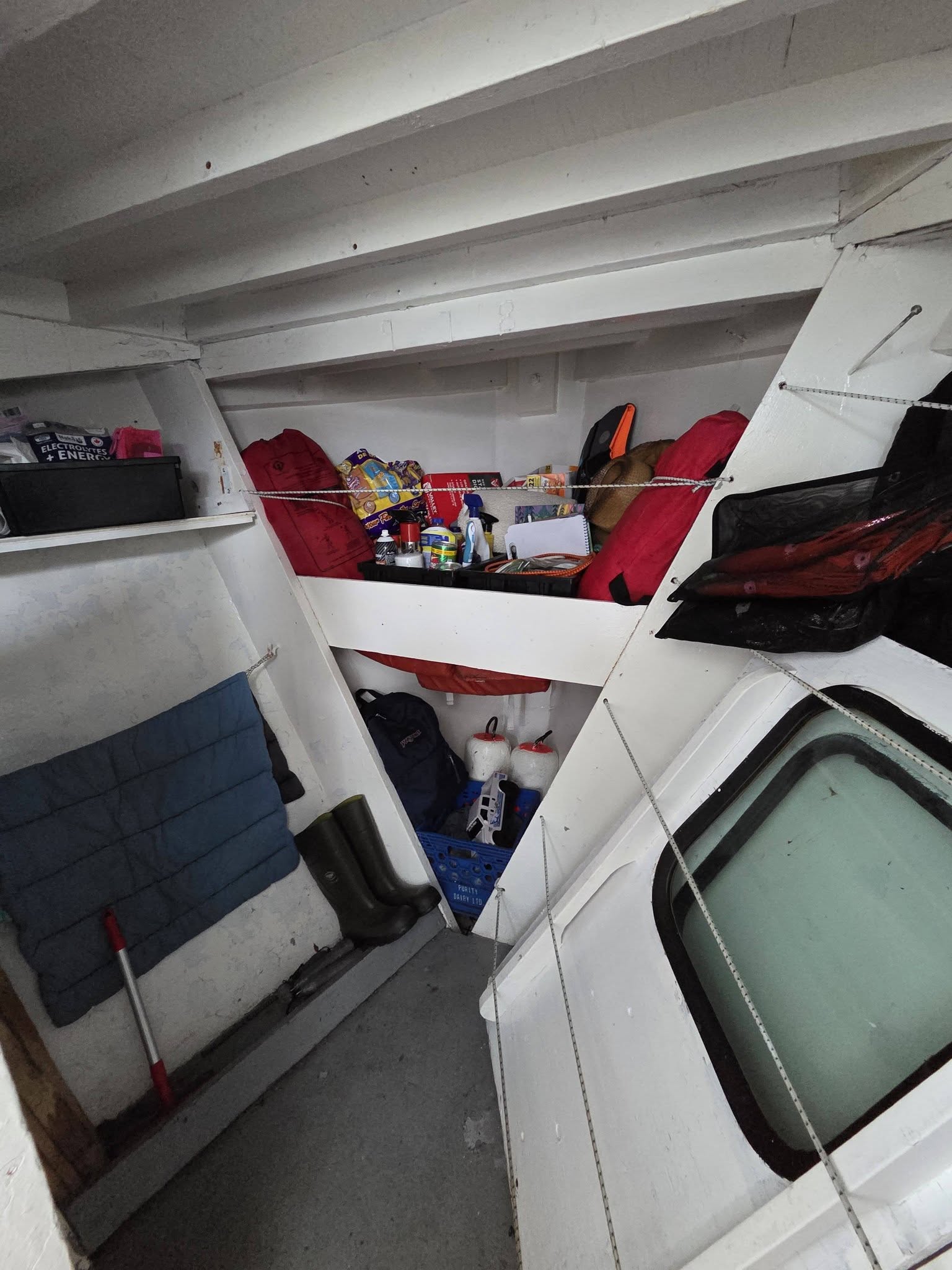A narrow, cluttered interior space with white walls and ceiling beams, containing shelves, a blue quilted mat, a broom, and various packed items including red and orange bags and boxes.
