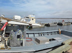 A person in a gray work jacket stands on the deck of a metal boat docked at a harbor, with other boats and water visible in the background under a cloudy sky.