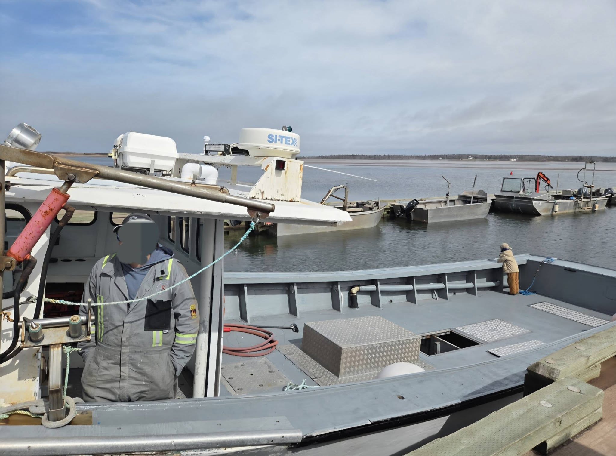 A person in a gray work jacket stands on the deck of a metal boat docked at a harbor, with other boats and water visible in the background under a cloudy sky.