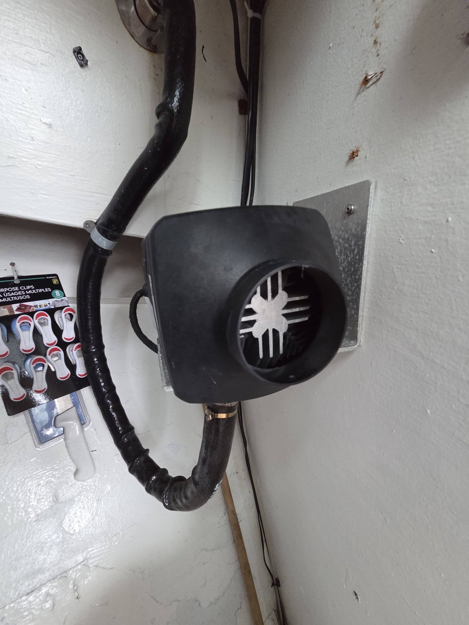 A black marine electric fan mounted on a white wall, with its black intake hose connected and a white propeller visible inside the fan opening.