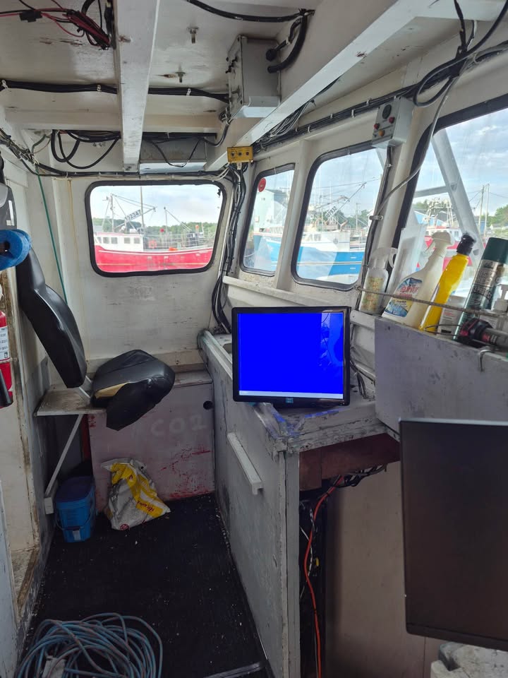 The interior of a boat cabin with a black steering wheel, a computer monitor displaying a solid blue screen on a console table, coiled cables, and windows looking out onto water and other boats.