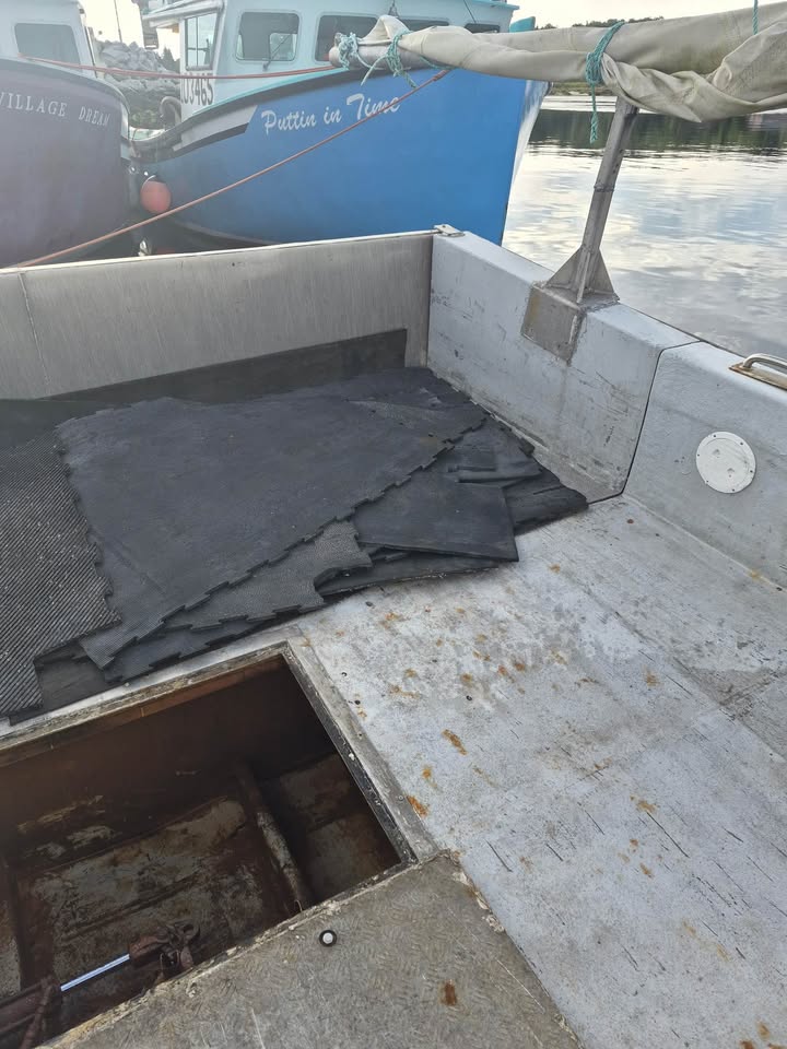 A set of dark gray to black textured mats or floor liners laid out inside the deck of a boat, with water and other boats visible in the background.