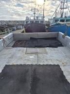 Black interlocking foam mats laid on the deck of a boat in a harbor, with other boats and a rocky breakwater visible in the background.