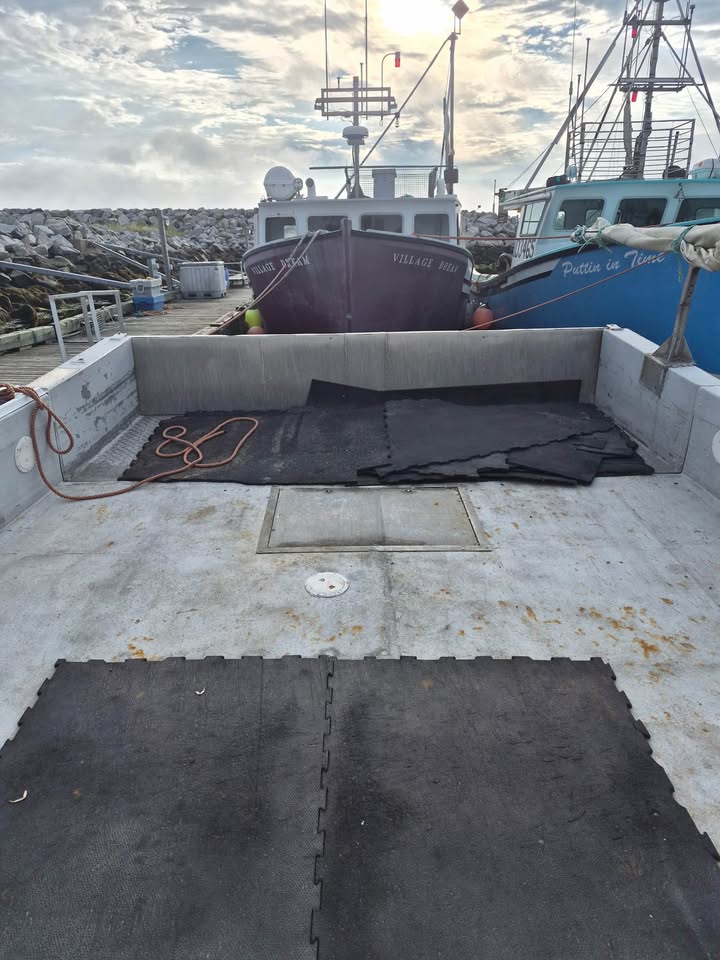 Black interlocking foam mats laid on the deck of a boat in a harbor, with other boats and a rocky breakwater visible in the background.