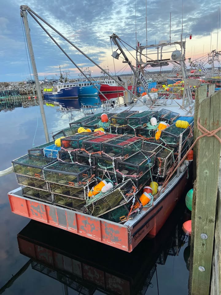 A red fishing boat docked at a harbor, loaded with stacked lobster traps and colorful buoys, with other boats and a cloudy sky in the background.