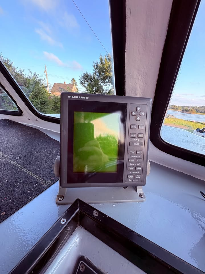 A Fuguro brand marine radar screen or navigation display device mounted on a dashboard inside a boat cabin, with water and trees visible through the windows.