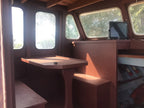The interior of a wooden boat cabin featuring a built-in table and bench, large arched windows with bright daylight, and a small monitor on the console.
