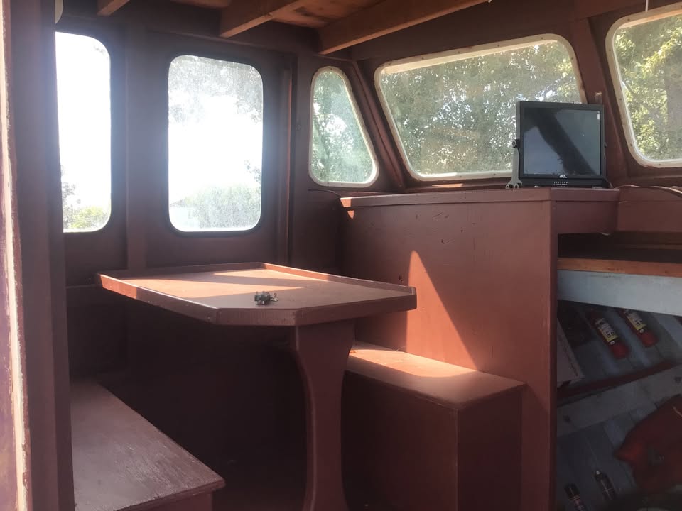 The interior of a wooden boat cabin featuring a built-in table and bench, large arched windows with bright daylight, and a small monitor on the console.