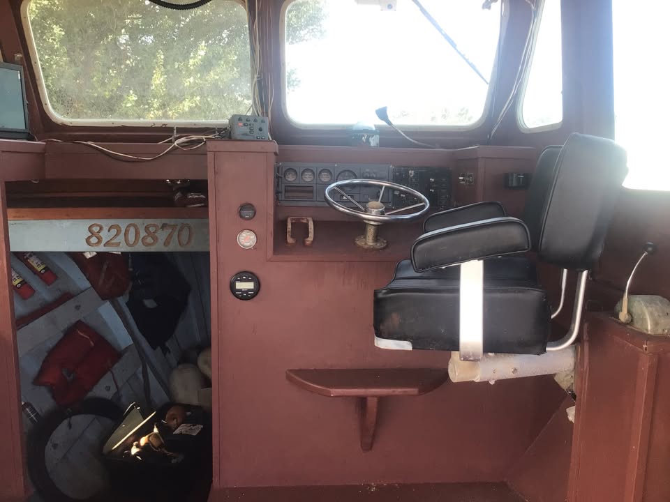 The interior of a boat cabin with a reddish-brown wooden dashboard and console, a black captain's seat with a white support frame, a chrome steering wheel, and windows showing bright outdoor light.
